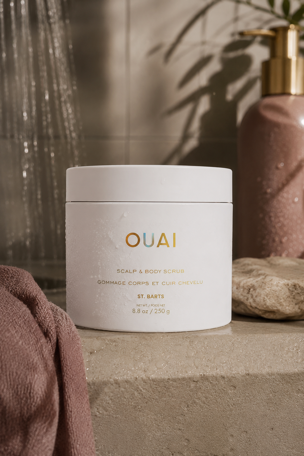 OUAI St. Barts Scalp & Body Scrub displayed on a warm stone ledge with a mauve towel in a softly lit bathroom.